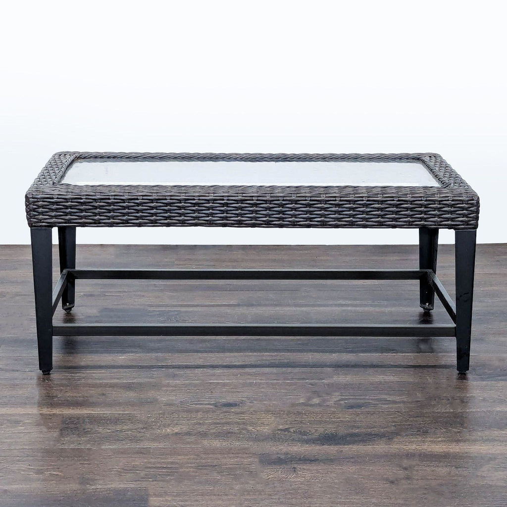 Rectangular table with metal frame, wicker border, and glass top, suitable for indoor or outdoor spaces.  