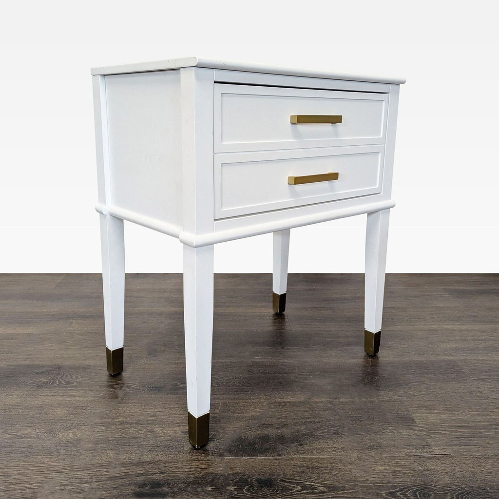 3. Angled view of a white end table with two drawers and gold accents on handles and leg tips, on dark floor.
