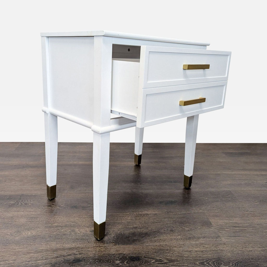 2. Side view of white end table featuring two drawers, gold handles, and gold-capped legs, one drawer open.