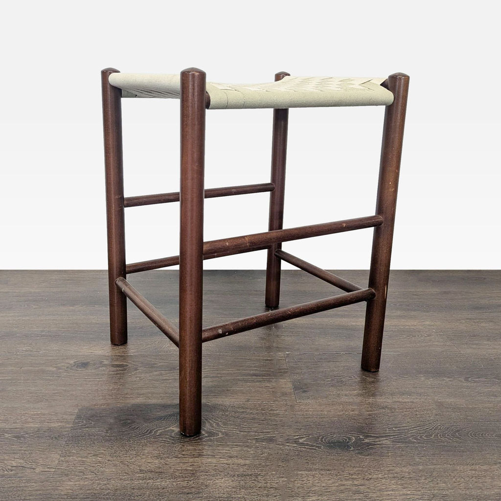 1. Wooden stool with round legs and a woven light fabric seat, placed on a wood floor, angled view.  