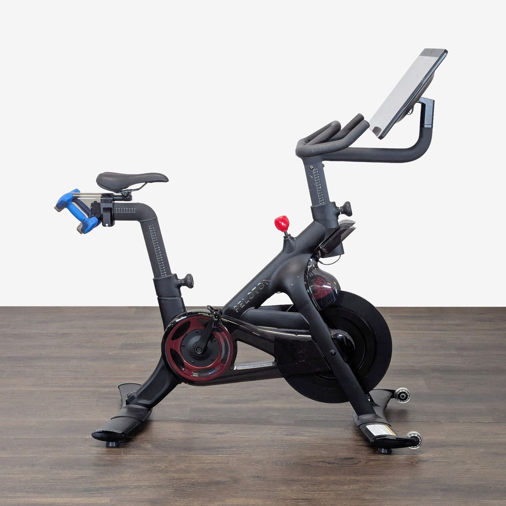 Peloton exercise bike with touchscreen, blue dumbbells, and adjustable seat for interactive home workouts.