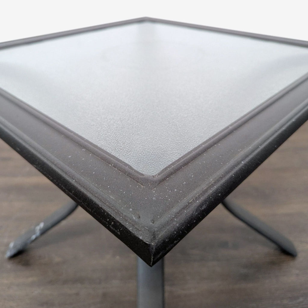 3. Close-up of a glass-topped square table with a sleek metal edge and sturdy legs, ideal as a side or patio table.