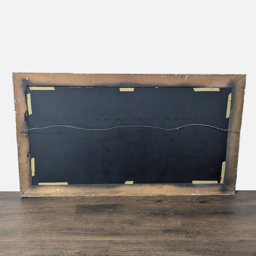 The back of a rectangular mirror with a wooden frame, metal hanging wire, and taped edges is shown.  
