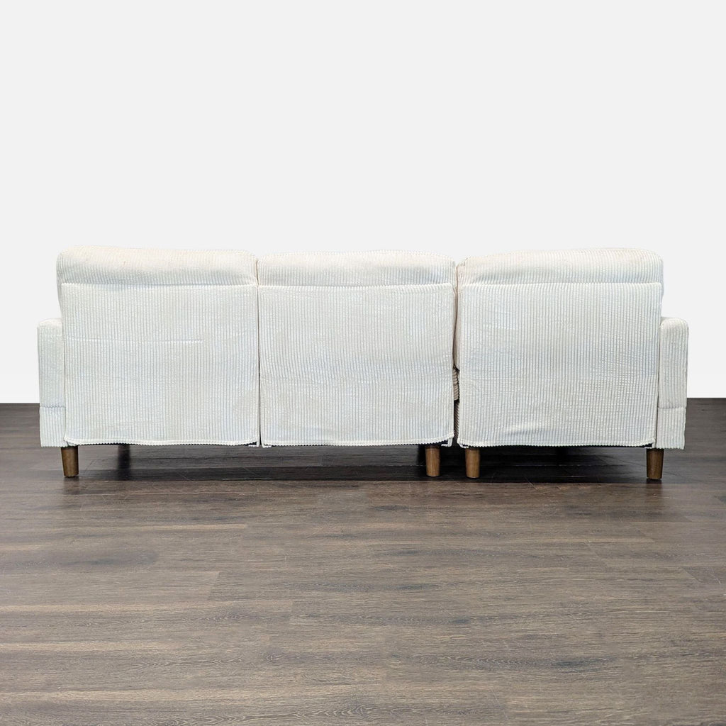 Rear view of a three-seat light beige corduroy sectional with sturdy wooden legs, shown on a dark floor.