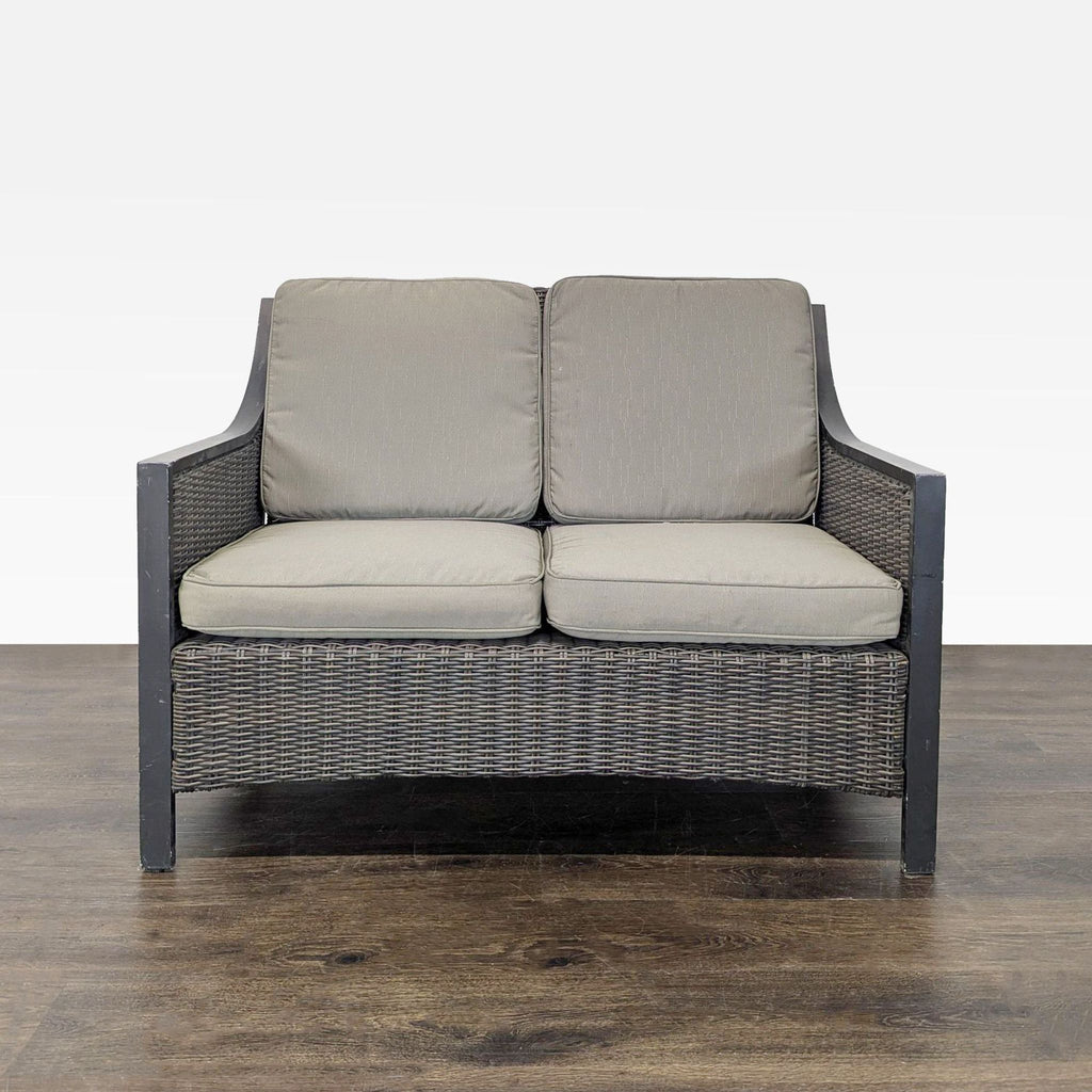 Outdoor loveseat with dark woven wicker frame and thick gray cushions, suitable for patios or gardens.