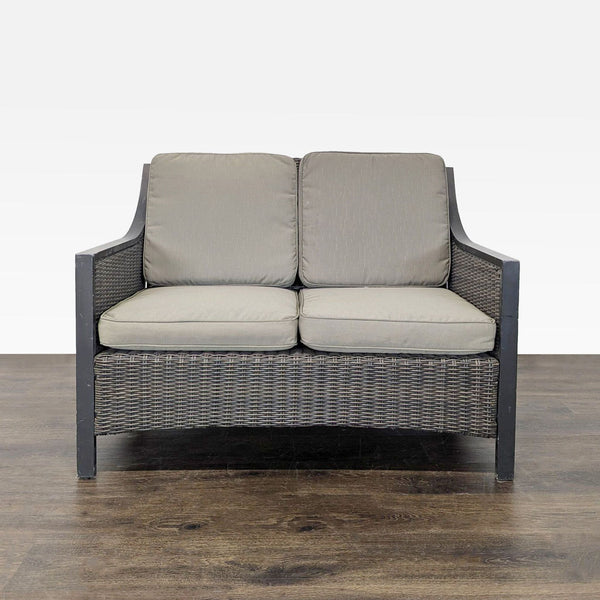 Outdoor loveseat with dark woven wicker frame and thick gray cushions, suitable for patios or gardens.