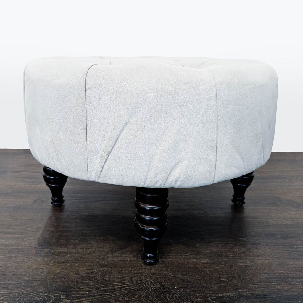 2. Side angle of a circular light grey ottoman with elegant black turned legs, on dark wood flooring.