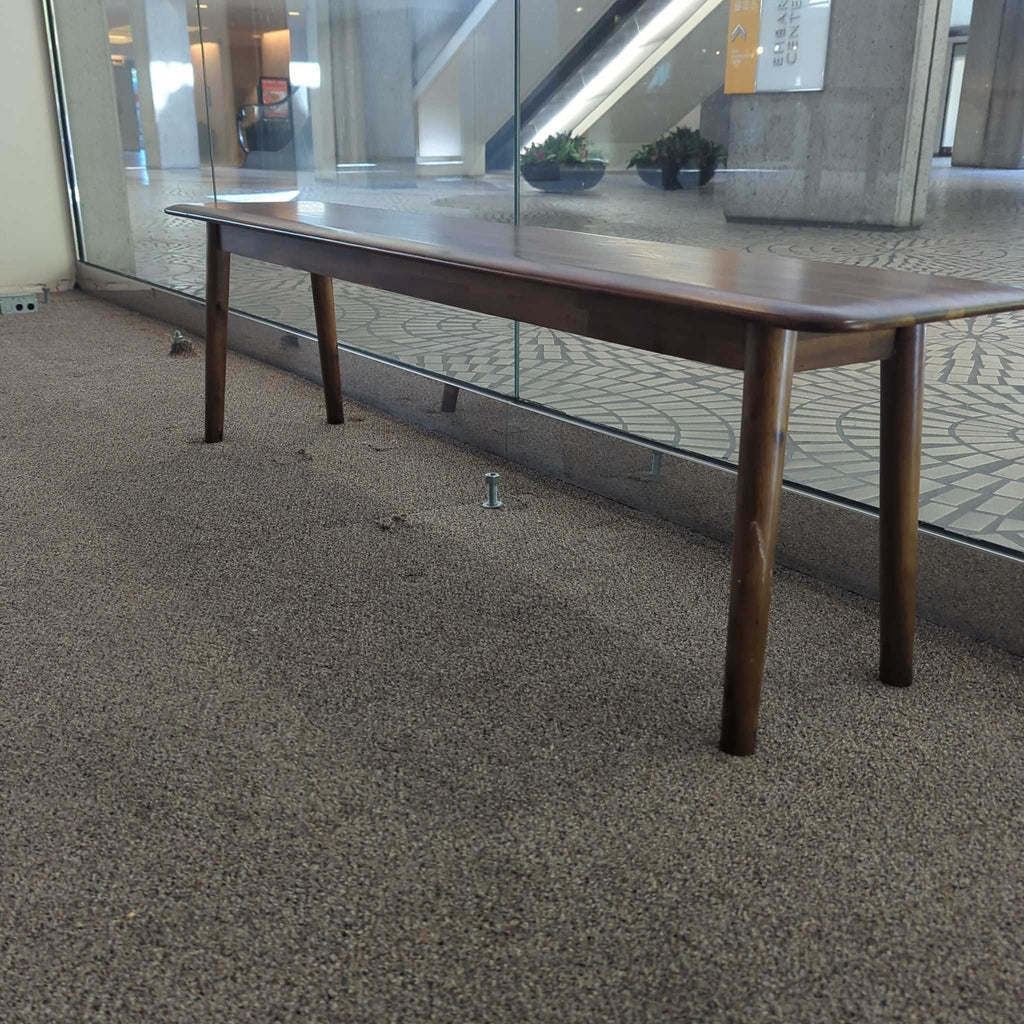2. Side view of a sleek wooden EQ3 bench with tapered legs, set against a large window in a modern space.