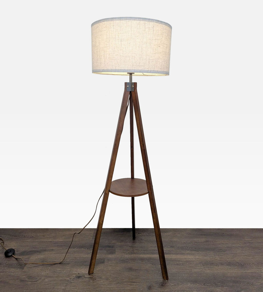 Modern tripod floor lamp with a wooden base, built-in shelf, and fabric shade for soft lighting.