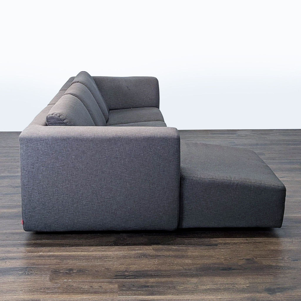 3. Side view of a dark grey EQ3 sectional sofa with a chaise, on a wooden floor, minimalist style.