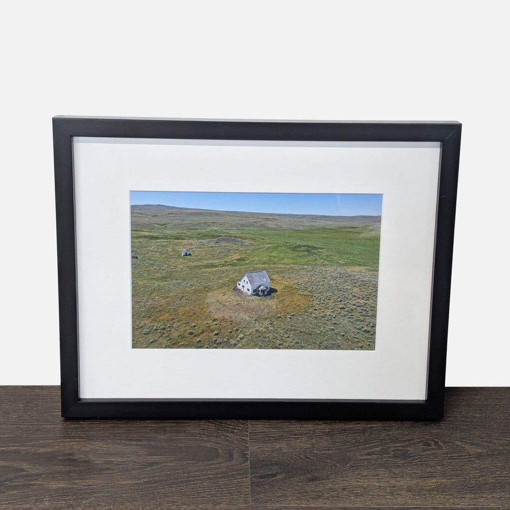 Aerial print of an old farmhouse in a grassy field, framed in black with a white mat, perfect for wall display.