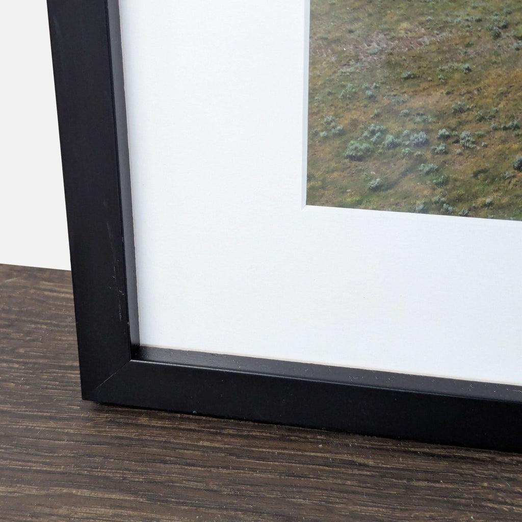 Print depicting a wide grassy field and vintage farmhouse, elegantly presented with a simple black frame and mat.