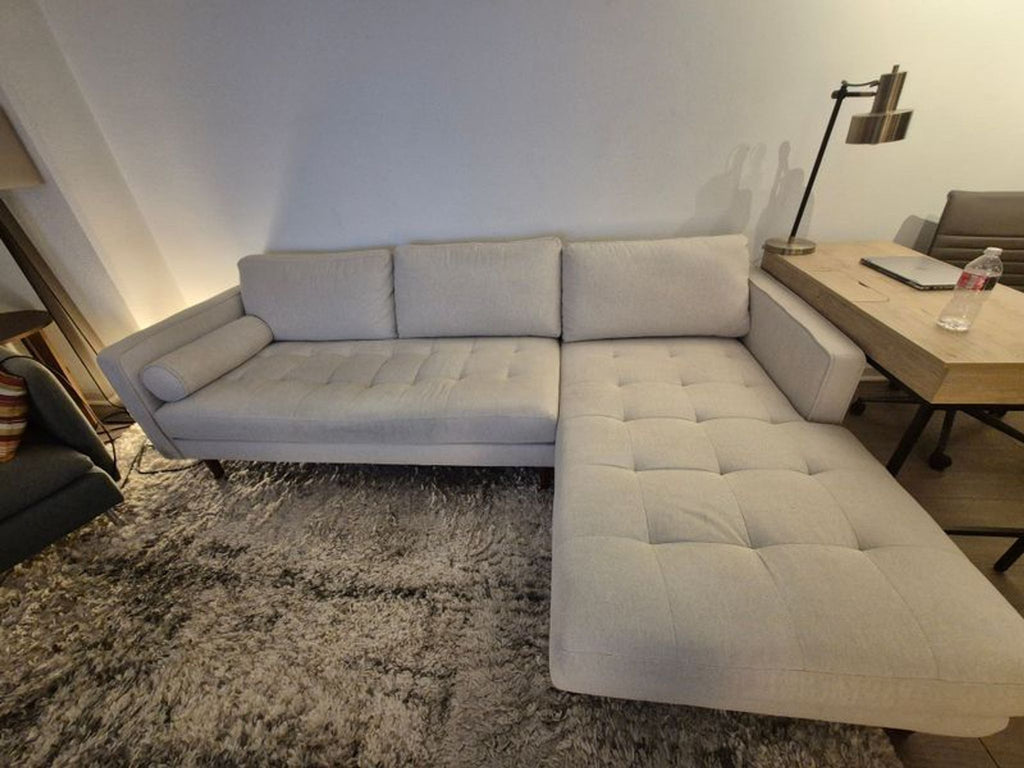 2. Modern LUKAS OPTICAL sectional sofa in light fabric with chaise lounge, placed beside a wooden desk and lamp.