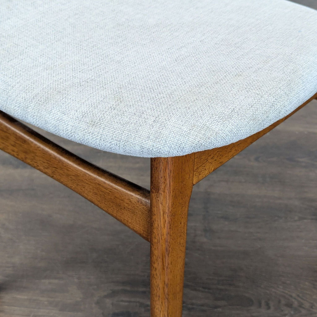 2. Close-up of the light fabric seat cushion and wooden leg of a West Elm dining chair on a dark wood floor.