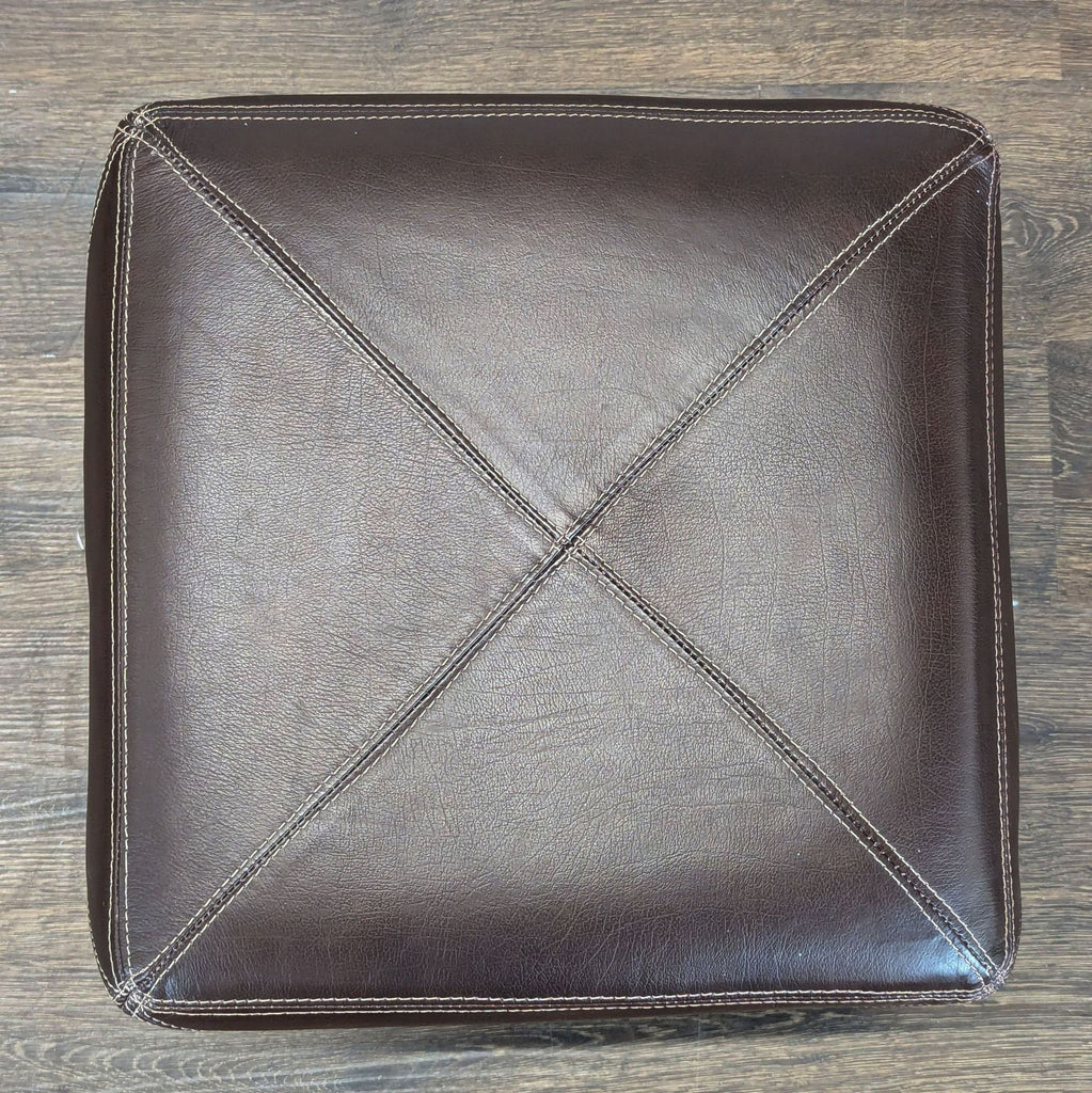 1. Top view of a square, dark brown leather ottoman with visible stitched seams and X pattern.