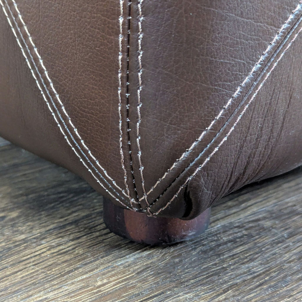 3. Bottom corner of a brown leather ottoman showing detailed stitching and a short wooden leg on wood flooring.