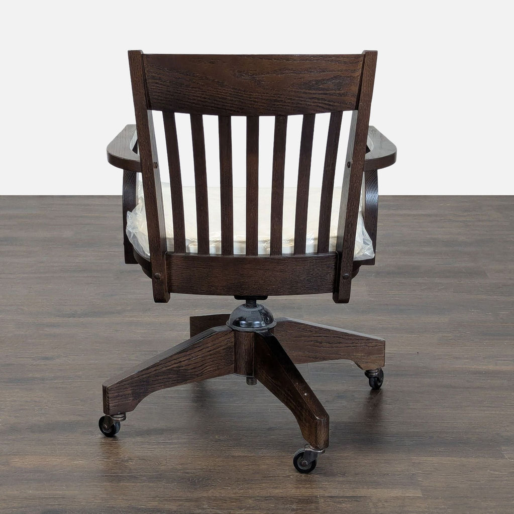 1. Rear view of a dark wood Pottery Barn office chair with slatted back and wheels on wood floor.