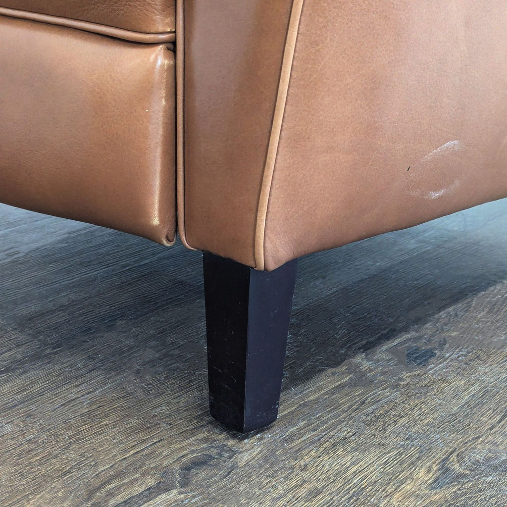 3. Detail of the bottom corner and dark wooden leg of a Room & Board cognac leather lounge chair on wood flooring.