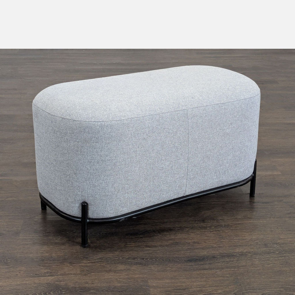 1. Modern gray upholstered ottoman with black metal legs, placed on dark wood flooring, side view.