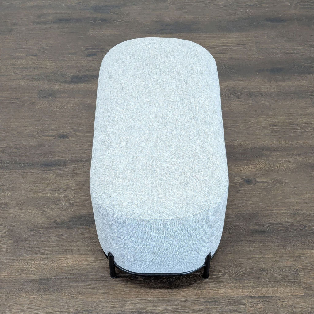 2. Top-down view of an oval-shaped, light gray fabric ottoman with a sturdy black metal base.