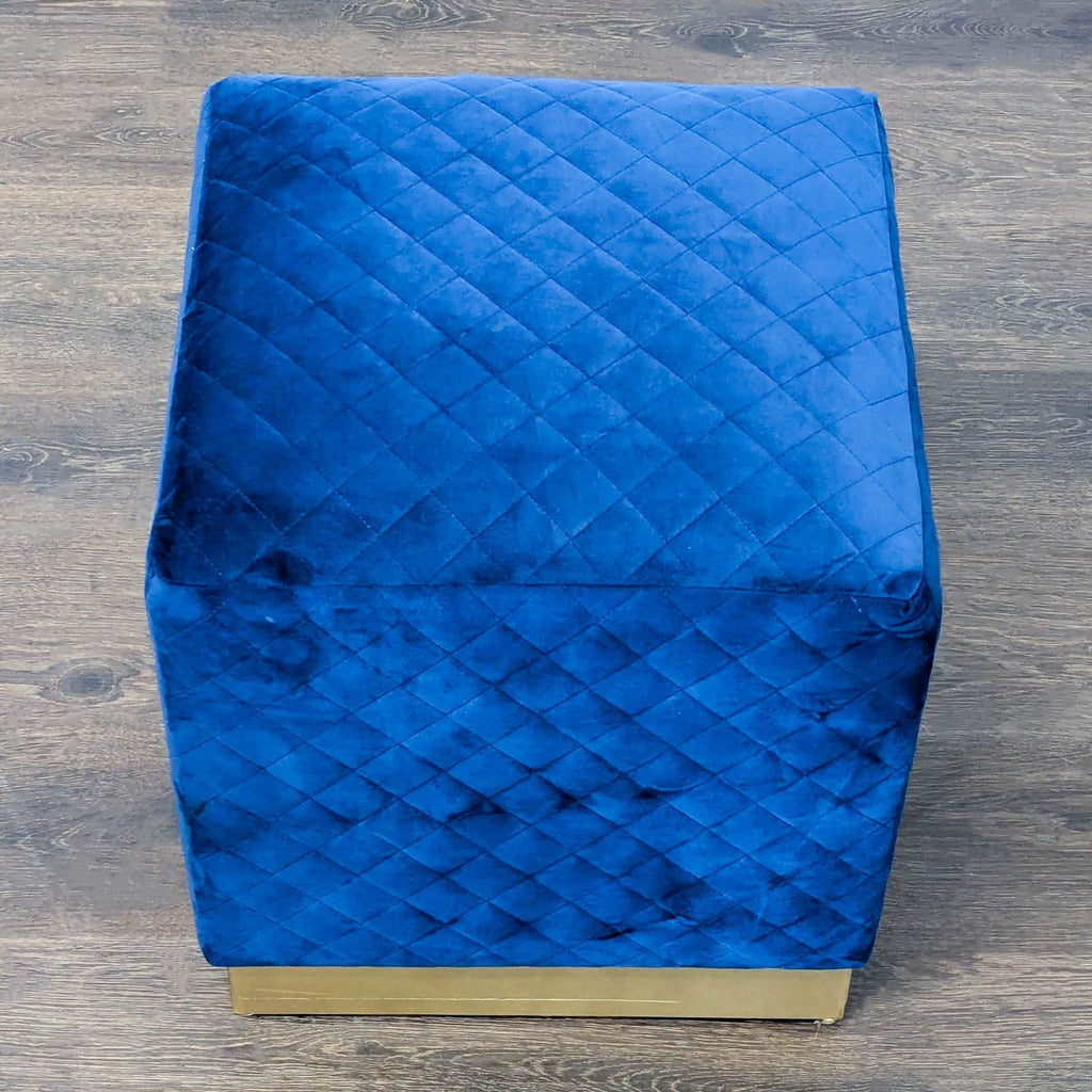 Chic upholstered ottoman in blue velvet with quilted top and shiny gold base for versatile seating.