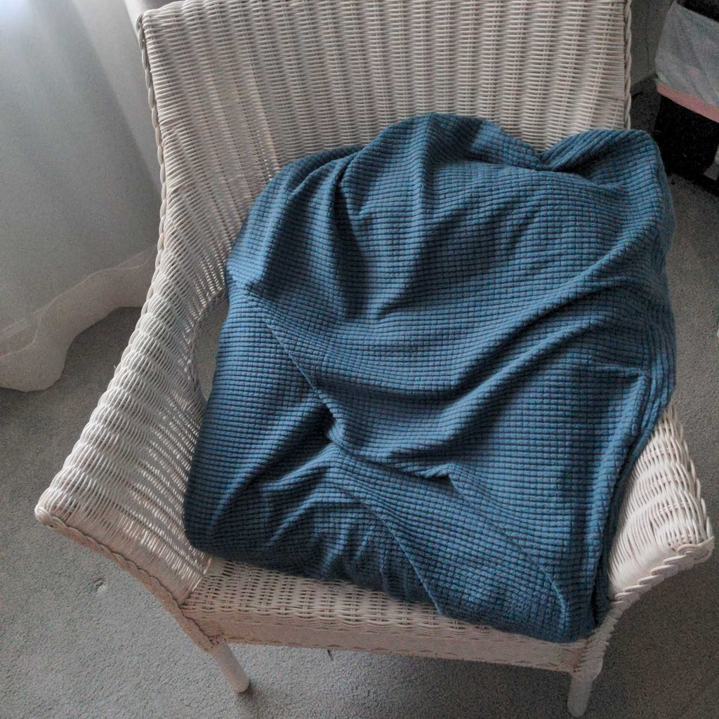 1. White wicker lounge chair with a blue textured blanket draped over the seat, next to a window.