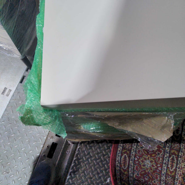 1. Close-up of a white West Elm end table corner, partly wrapped in green bubble wrap and plastic.