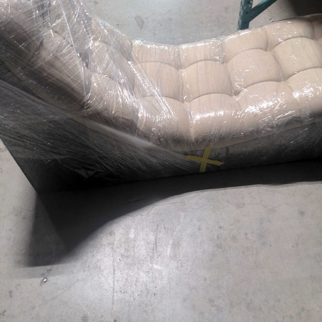 Beige tufted lounge chair wrapped in plastic, partially viewed from the side on a warehouse floor.  