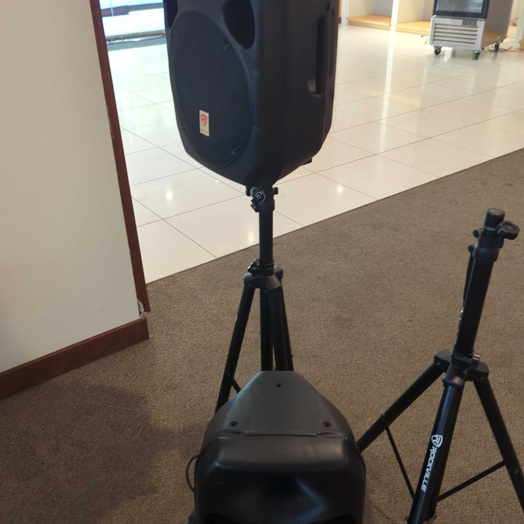 3. Rockville brand audio speaker mounted on a tripod, next to another stand, on brown carpet and tile flooring.