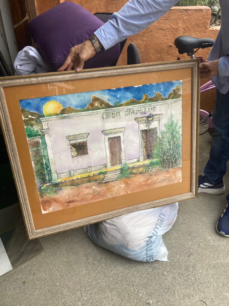 2. Two people hold a framed Reperch painting showing a rustic house in a mountainous landscape.