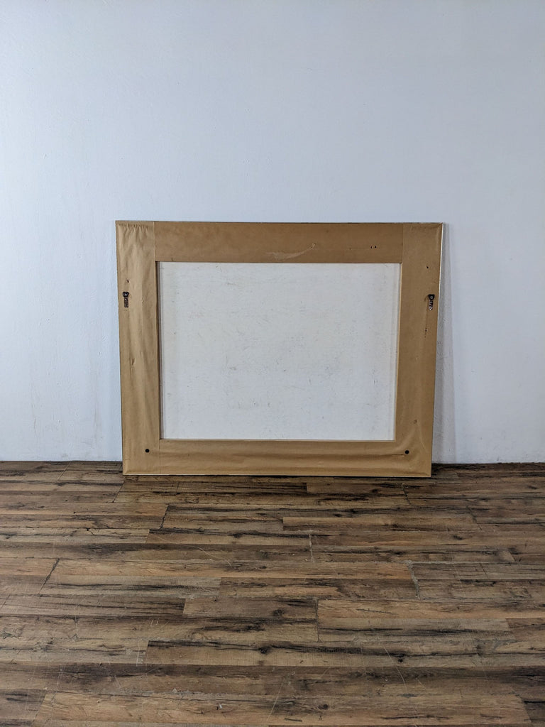 Back of a framed painting showing the wooden frame and canvas against a wood floor.
