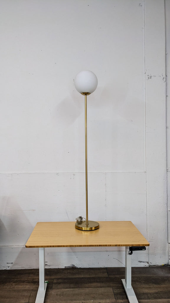 Modern floor lamp by Reperch with a brass finish and white globe shade, set on a wooden table.