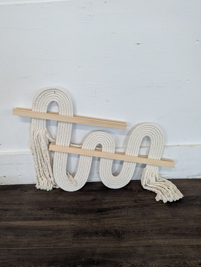 2. Sculptural rope fiber artwork with natural wood bars and twisted fringe, leaning against a white wall and dark floor.