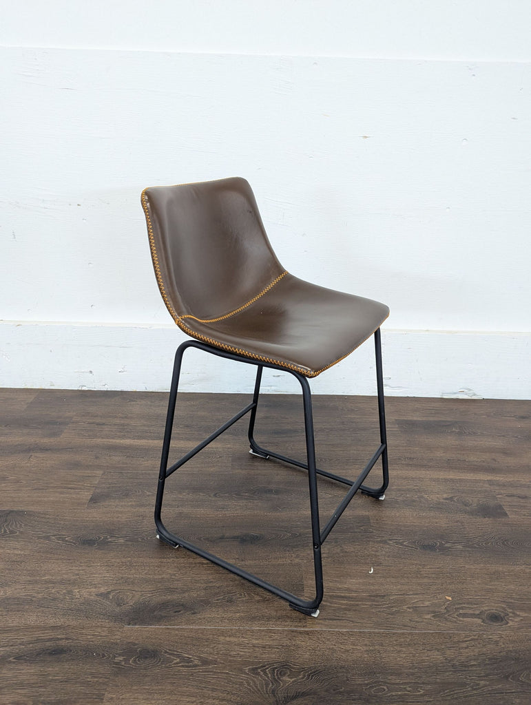 3. Side view of a Reperch brown leather bar stool with metal legs, positioned on a dark wood floor and white wall background.