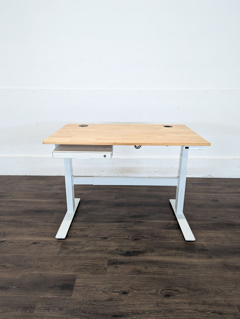 3. Compact Uplift adjustable desk with dual grommet holes and lockable drawer, standing on dark wood floors.