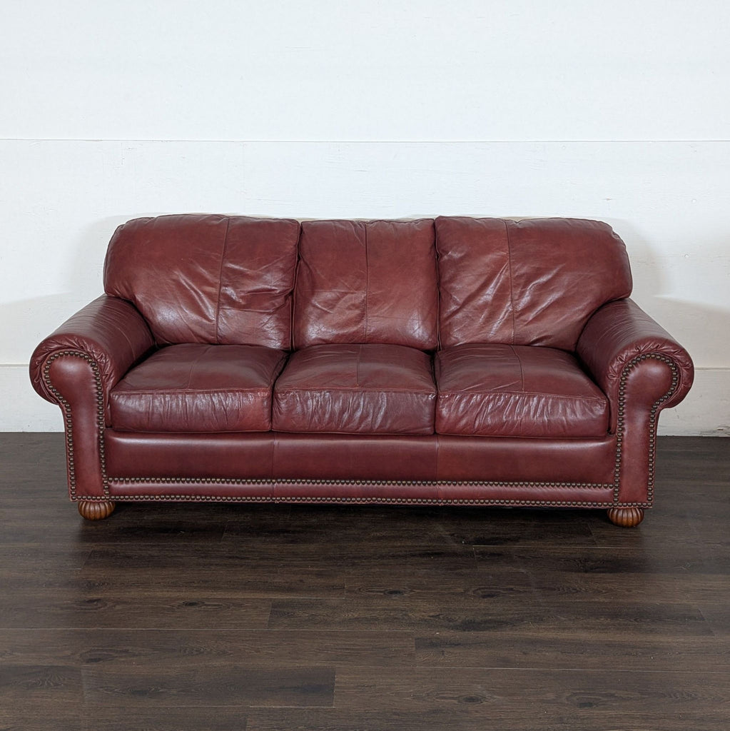 3. Reperch 3-seat maroon leather sofa with rolled arms and studded trim on a dark wood floor against a plain wall.