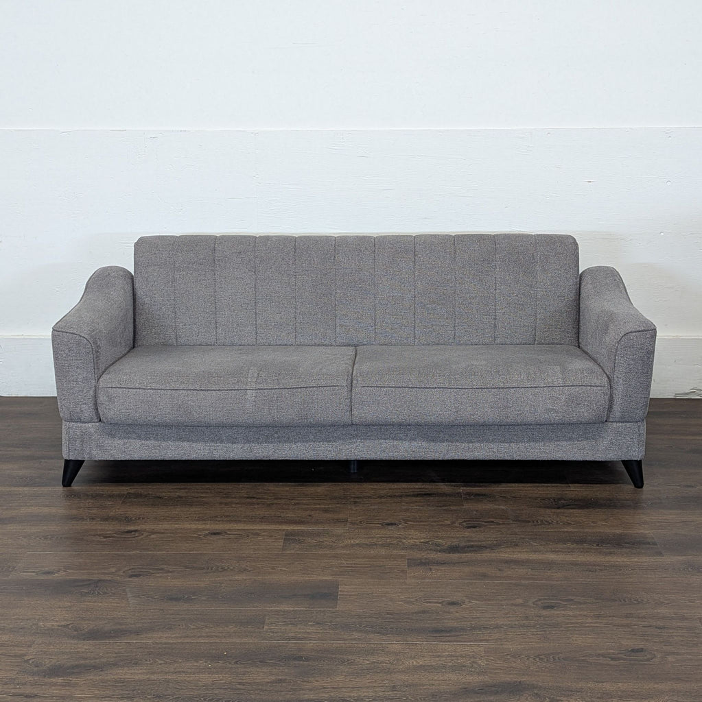 2. Gray upholstered 3-seat sofa with vertical stitching, seen from the front, on dark wooden flooring.