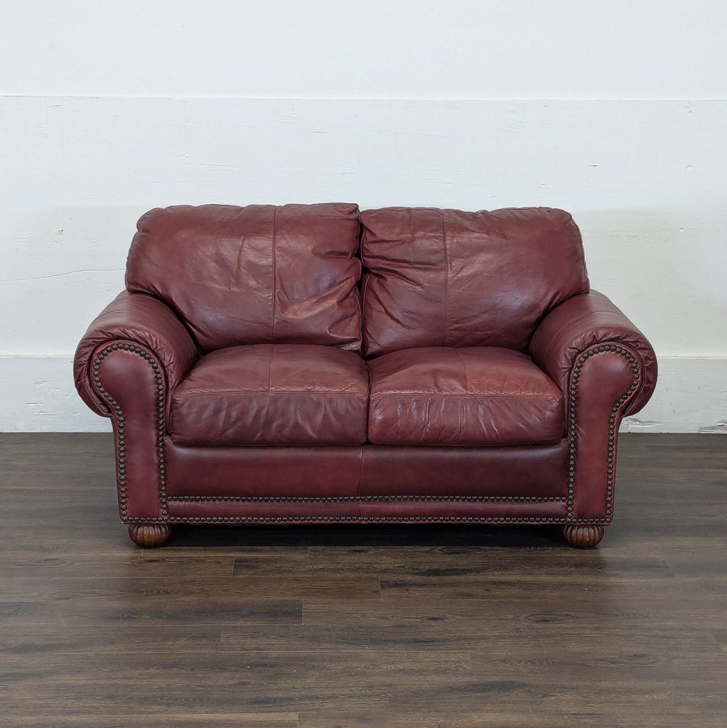 2. Maroon leather Reperch Loveseat with rolled arms and nailhead trim, set on a wooden floor against a white wall.
