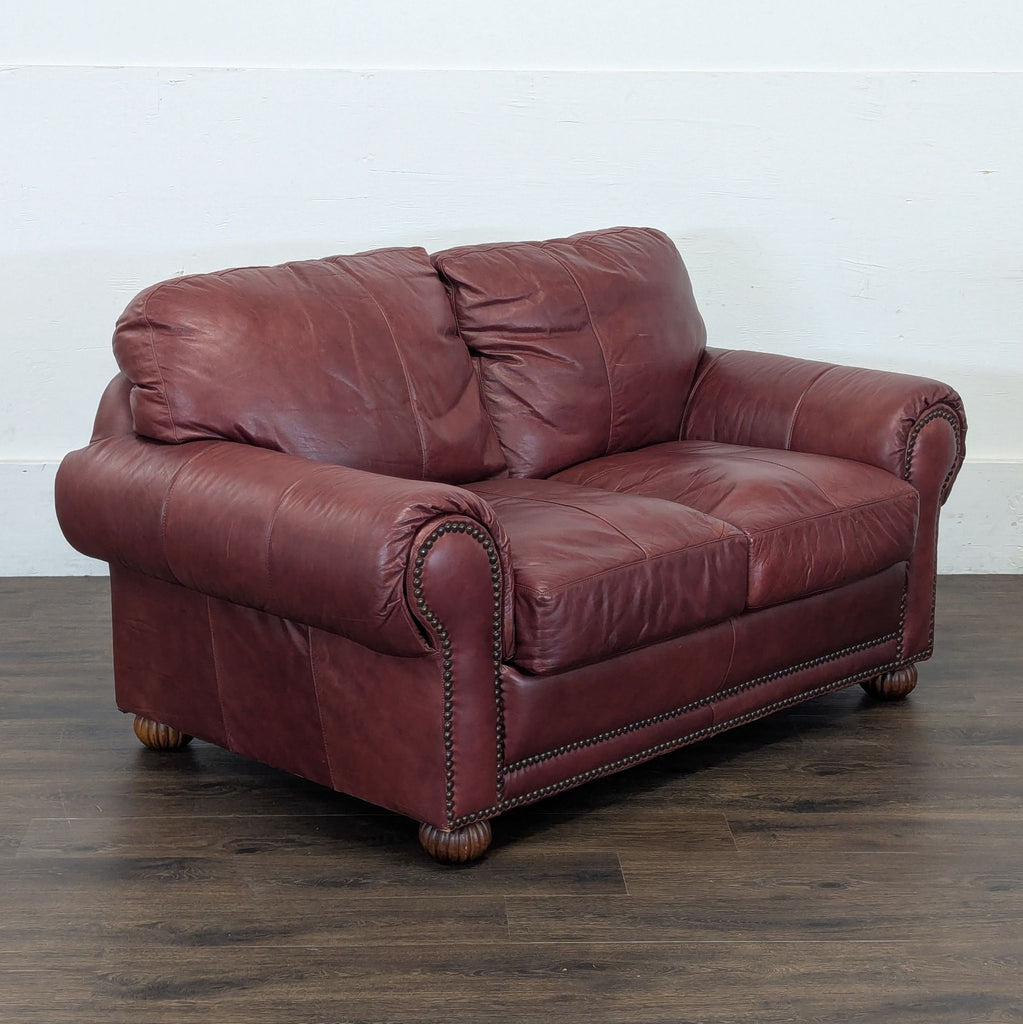3. Angled view of a maroon leather Reperch Loveseat with classic rolled arms and nailhead detailing on wood legs.