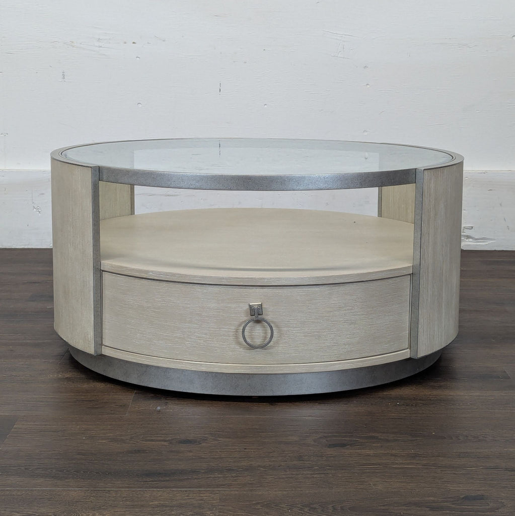2. Modern round coffee table, glass top, wood base with storage drawer and metallic trim, shown on dark wood floor.  