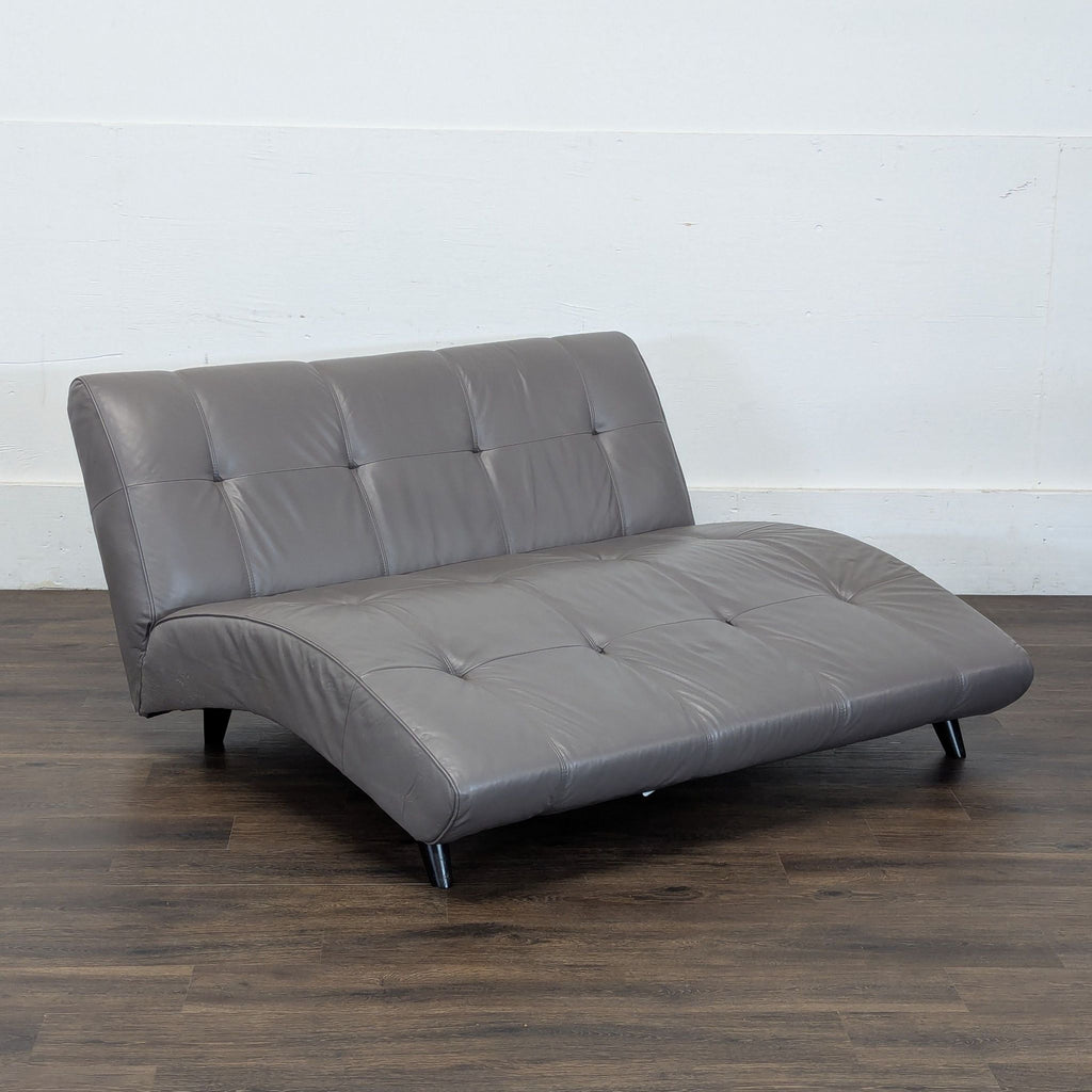 3. Angled view of a gray EQ3 tufted futon with curved seat, on a dark wood floor and plain background.