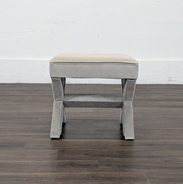 1. Reperch padded stool with grey velvet upholstery and crossed legs, viewed from the front, on wood flooring.