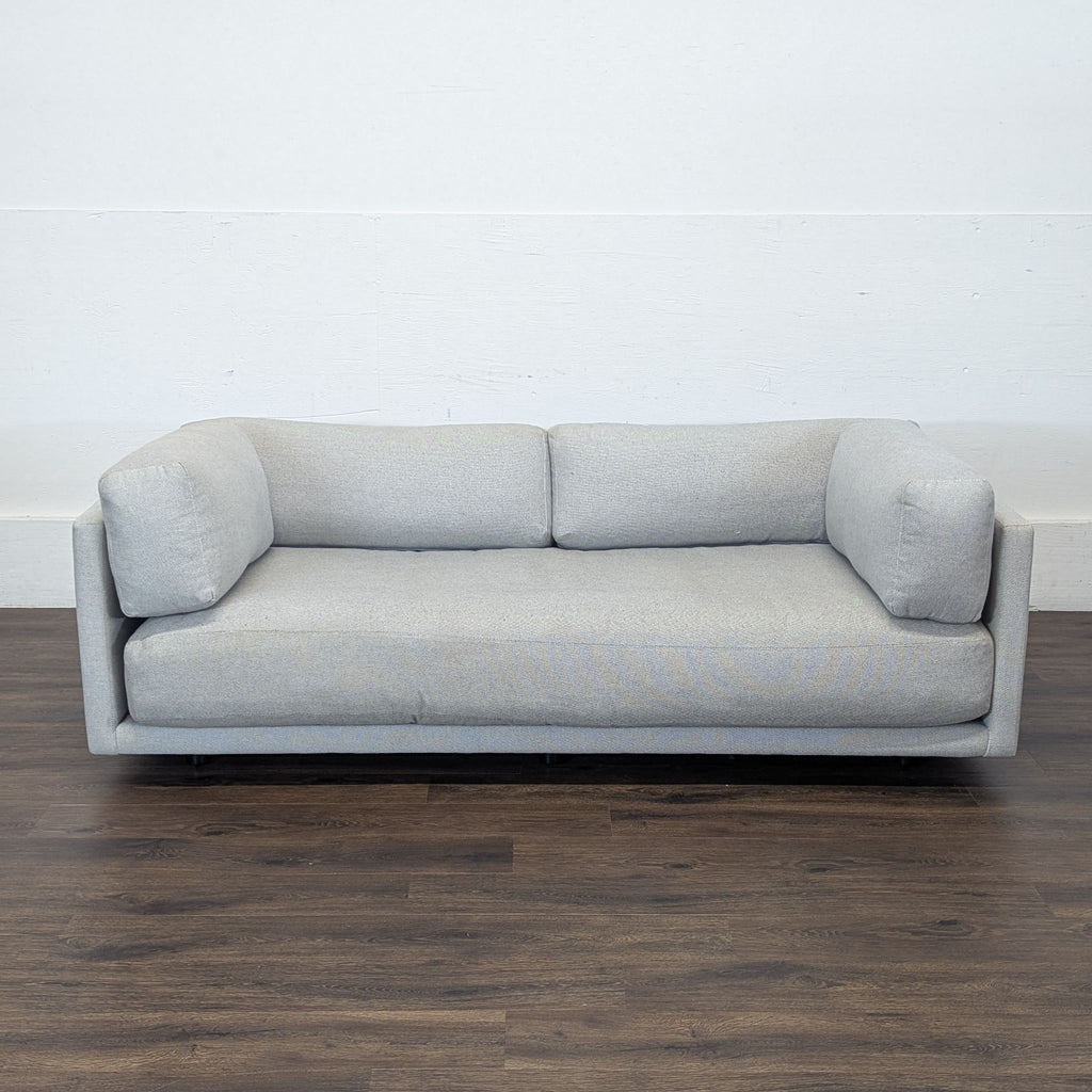 BluDot 3-seat sofa in soft gray fabric, shown against a white wall on dark wooden flooring.  