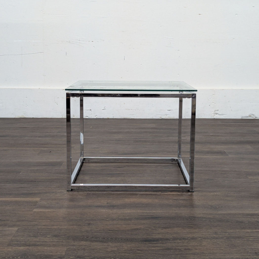 3. Minimalist glass and chrome end table with a square design against a white wall and wood flooring.