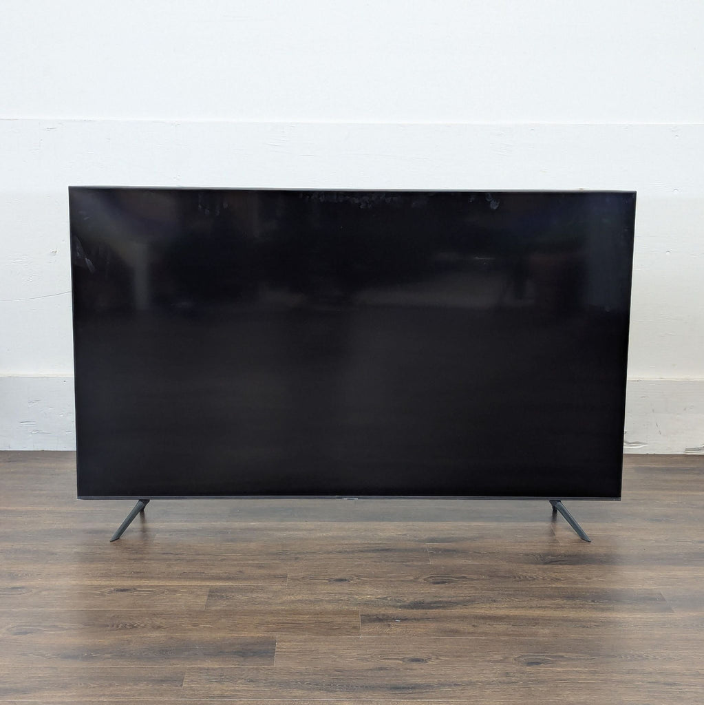 Front view of a Reperch TV with slim legs, placed on a wooden floor against a white wall, screen off.