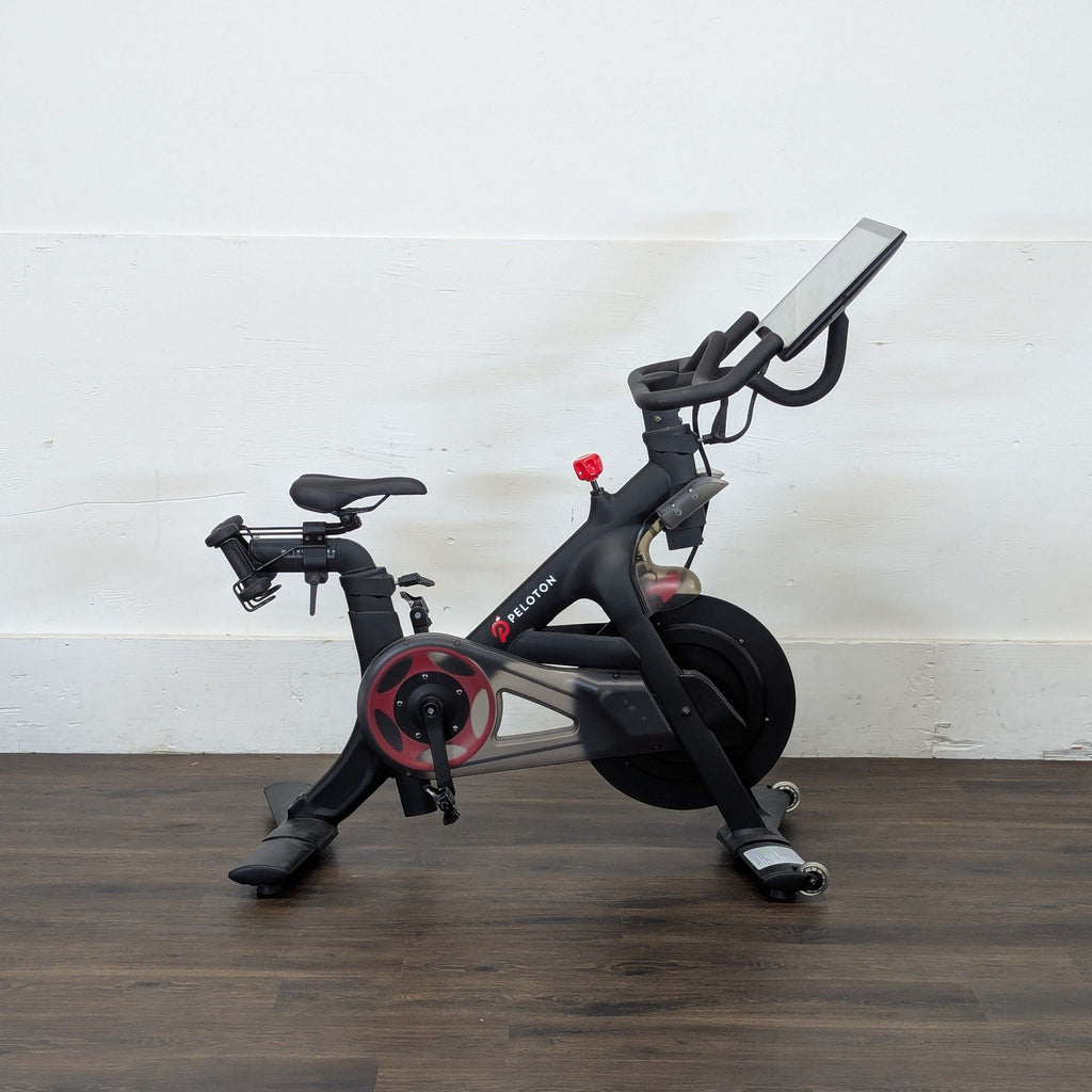 2. Side view of a Peleton gym bike with screen, photographed on a wooden floor against a white wall.