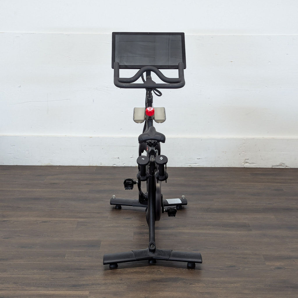 3. Front-facing Peleton exercise bike showing handlebars, seat, and large display, on a wood floor background.