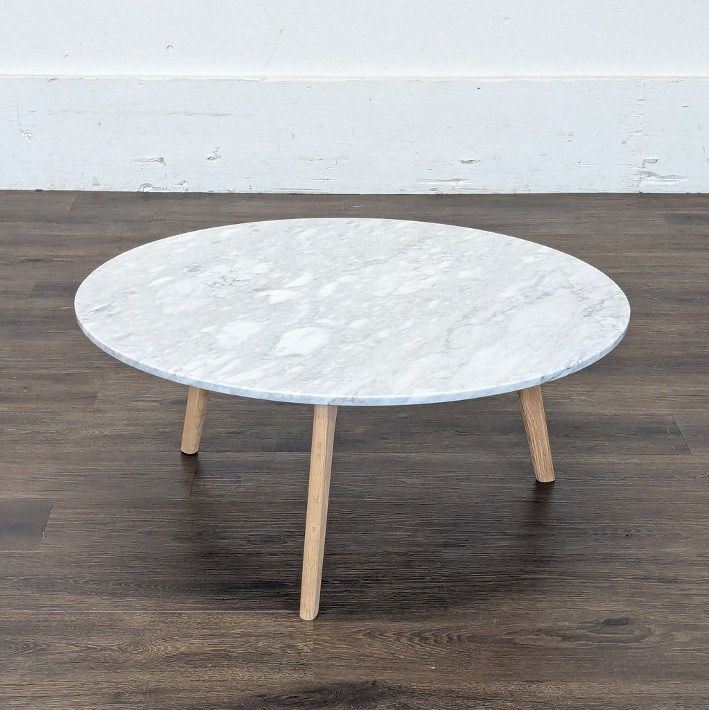 3. Circular marble coffee table by Article featuring tapered wood legs, set against a plain white wall and wood floor.