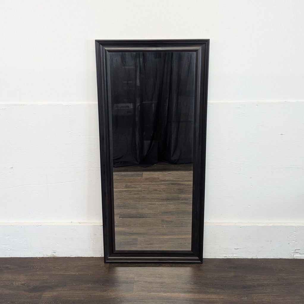 2. Ikea mirror in a dark frame placed against a white wall, reflecting wood flooring and a black curtain.