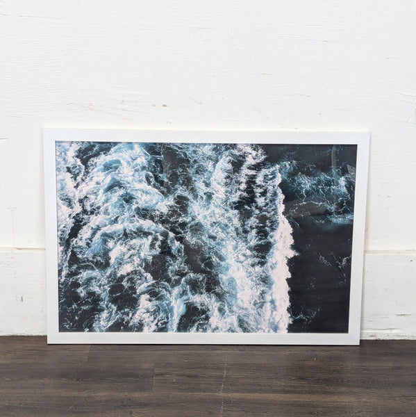 A framed print featuring an aerial view of ocean waves with a white frame, leaning against a white wall.  
